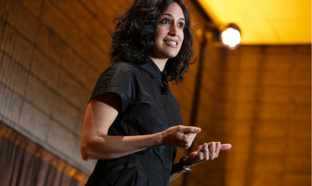 A speaker presenting on stage, engaging with an audience. She has long, dark hair and gestures expressively with her hands. The backdrop features a textured wall, and a spotlight illuminates her as she speaks, creating an intimate atmosphere.