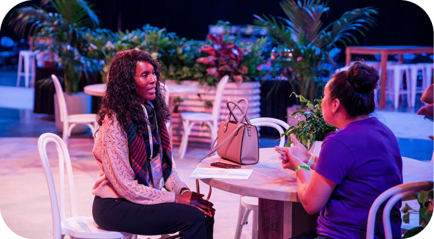 A woman with curly hair is sitting at a table and engaged in a conversation with another woman, who is wearing a purple shirt. They are surrounded by lush greenery and a few other tables in the background, indicative of an event setting. There is a beige handbag on the table in front of them, suggesting a casual yet professional atmosphere.