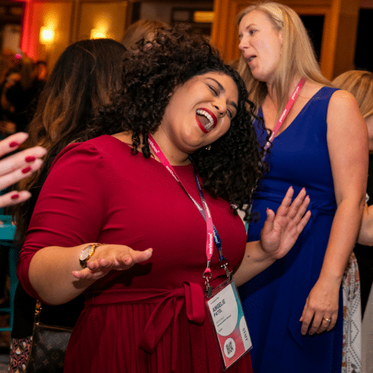 A woman in a red dress is joyfully dancing with her hands raised, surrounded by other attendees at a lively event. She has dark, curly hair and wears a name badge. Another woman in a blue dress smiles and appears to be singing nearby. The atmosphere is vibrant and celebratory.