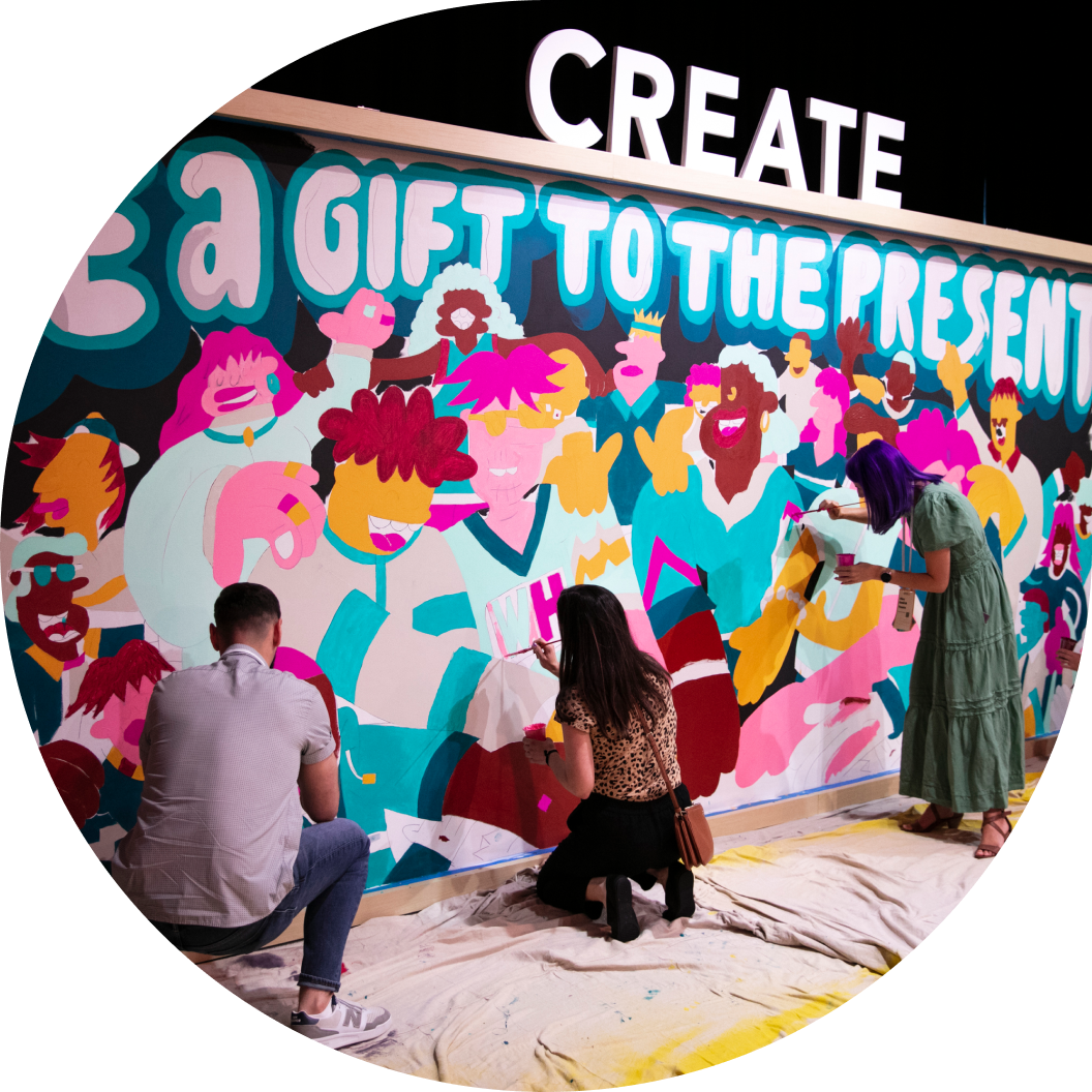 A colorful mural featuring diverse, stylized human figures is being painted by three individuals. In the foreground, two people are working on the mural; one is sitting and applying paint, while another is kneeling and focused on adding details. A large sign above the mural reads 