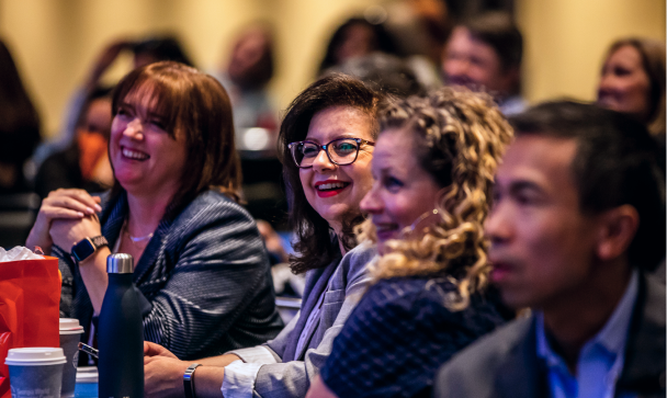 A group of smiling individuals engage attentively in a lively setting, likely during a conference or event. In the foreground, two women are prominently featured&mdash;one with glasses and the other with curly hair&mdash;while a man is visible on the right. The background shows several attendees, suggesting a larger audience. Various items such as cups and bags are placed on the table in front of them, contributing to the vibrant atmosphere.