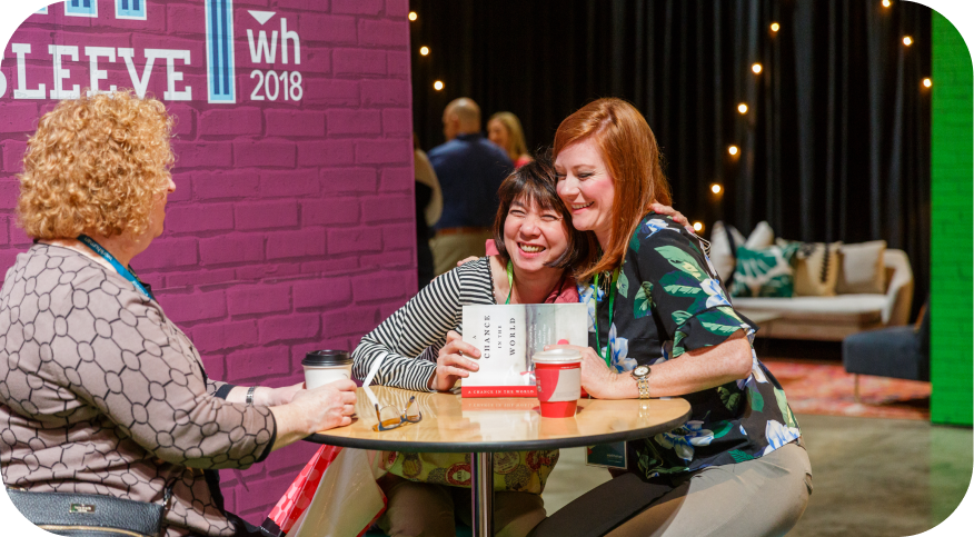 Two women smiling and embracing at a table, holding a book titled 