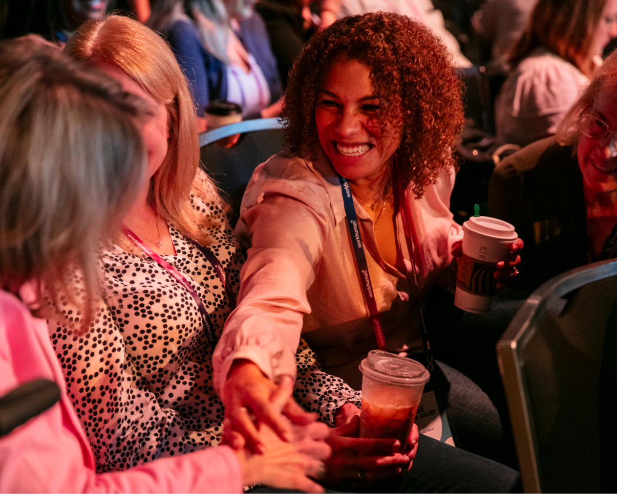 A joyful interaction among a group of attendees at a conference is depicted, with a woman in a light-colored shirt smiling broadly as she reaches out to shake hands with another participant. The other women, seated nearby, appear engaged and are holding beverages. The scene captures a warm, friendly atmosphere, highlighting connection and camaraderie among the attendees. The background features blurred silhouettes of additional participants, indicating a crowded event.