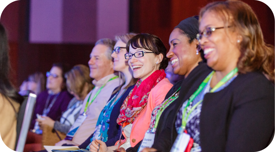 A diverse group of attendees seated in a conference setting, smiling and engaged. They are wearing name tags and have colorful lanyards. The atmosphere appears friendly and lively, indicating a positive experience at the event.
