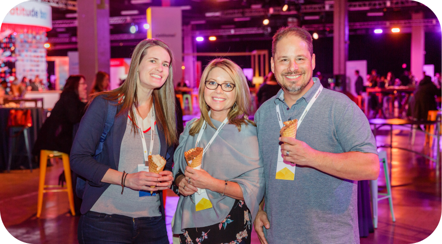 Three attendees at a Workhuman event are smiling and holding ice cream cones. They are gathered in a vibrant setting with colorful lighting and tables in the background, suggesting a lively atmosphere. All three are wearing event badges around their necks.