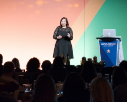 A woman in a black dress stands on stage speaking to an audience, with a brightly colored backdrop featuring shades of orange and green. A laptop is placed on a podium displaying the Workhuman logo. Attendees are seated in front, some visible in silhouette as they listen to the presentation.