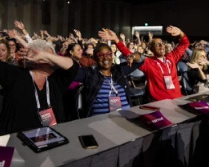 A lively audience at a conference with attendees engaged and raising their arms in celebration or response. In the foreground, three individuals are visible: one wearing glasses and smiling, another in a navy jacket, and a third in a red shirt. Tables in front of them display name tags and devices, capturing the energetic atmosphere of the event.