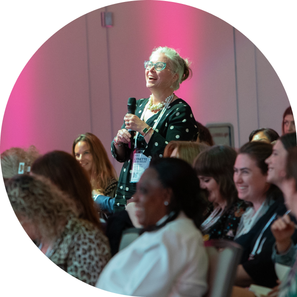 A woman with glasses and a polka dot blouse is standing in a crowd, smiling and holding a microphone as she engages during a conference. The audience around her is smiling, with a diverse group of attendees visible, all engaged in the moment. The background features soft pink lighting, creating a vibrant and inviting atmosphere.